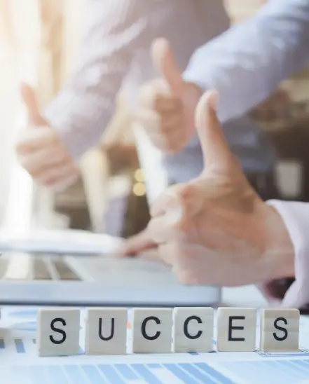 Three business professionals giving thumbs up behind wooden blocks spelling _SUCCESS_ on a desk with charts and documents