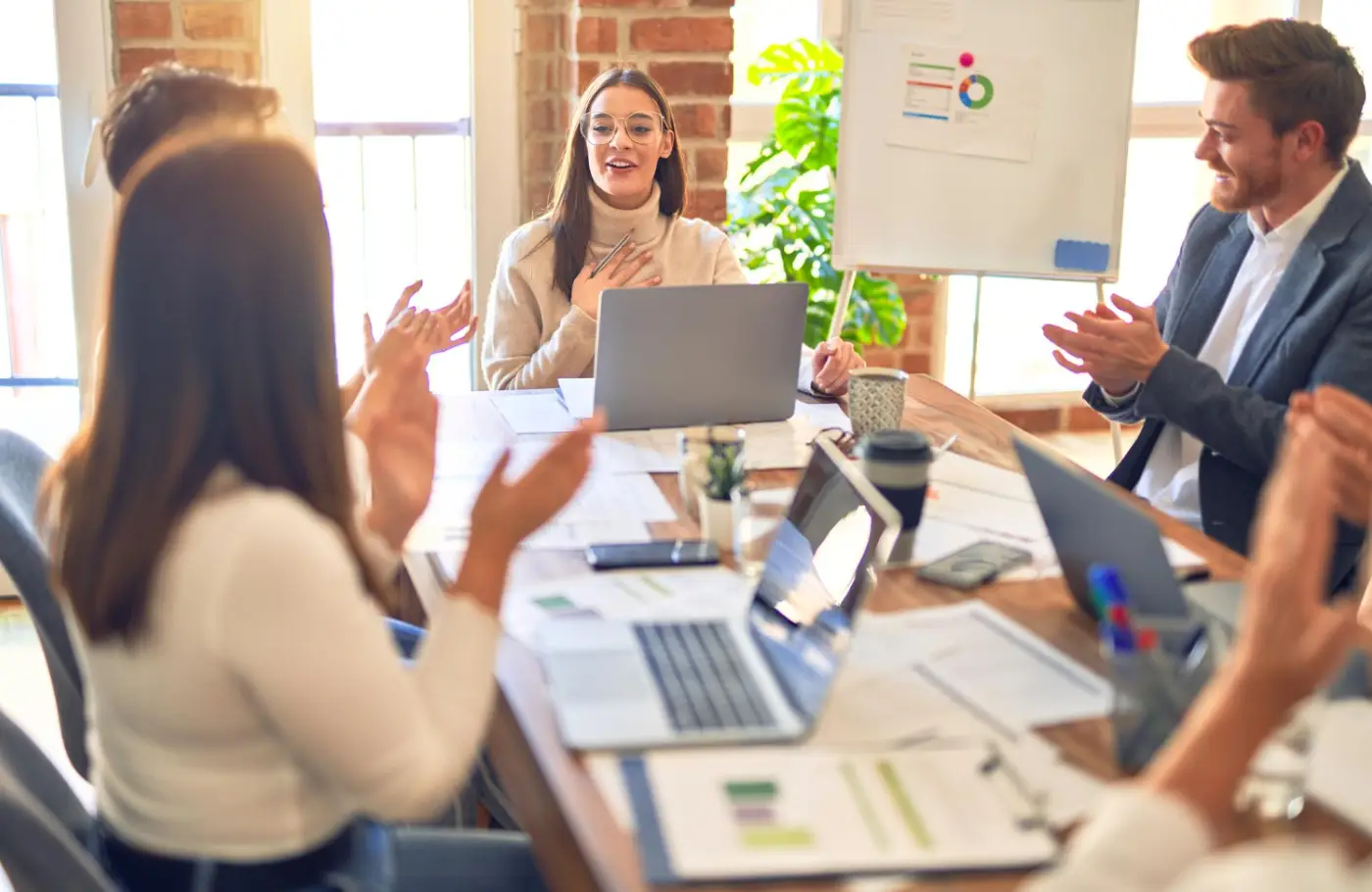 A group of professionals engaged in a meeting, clapping and discussing strategies, with laptops and documents on the table